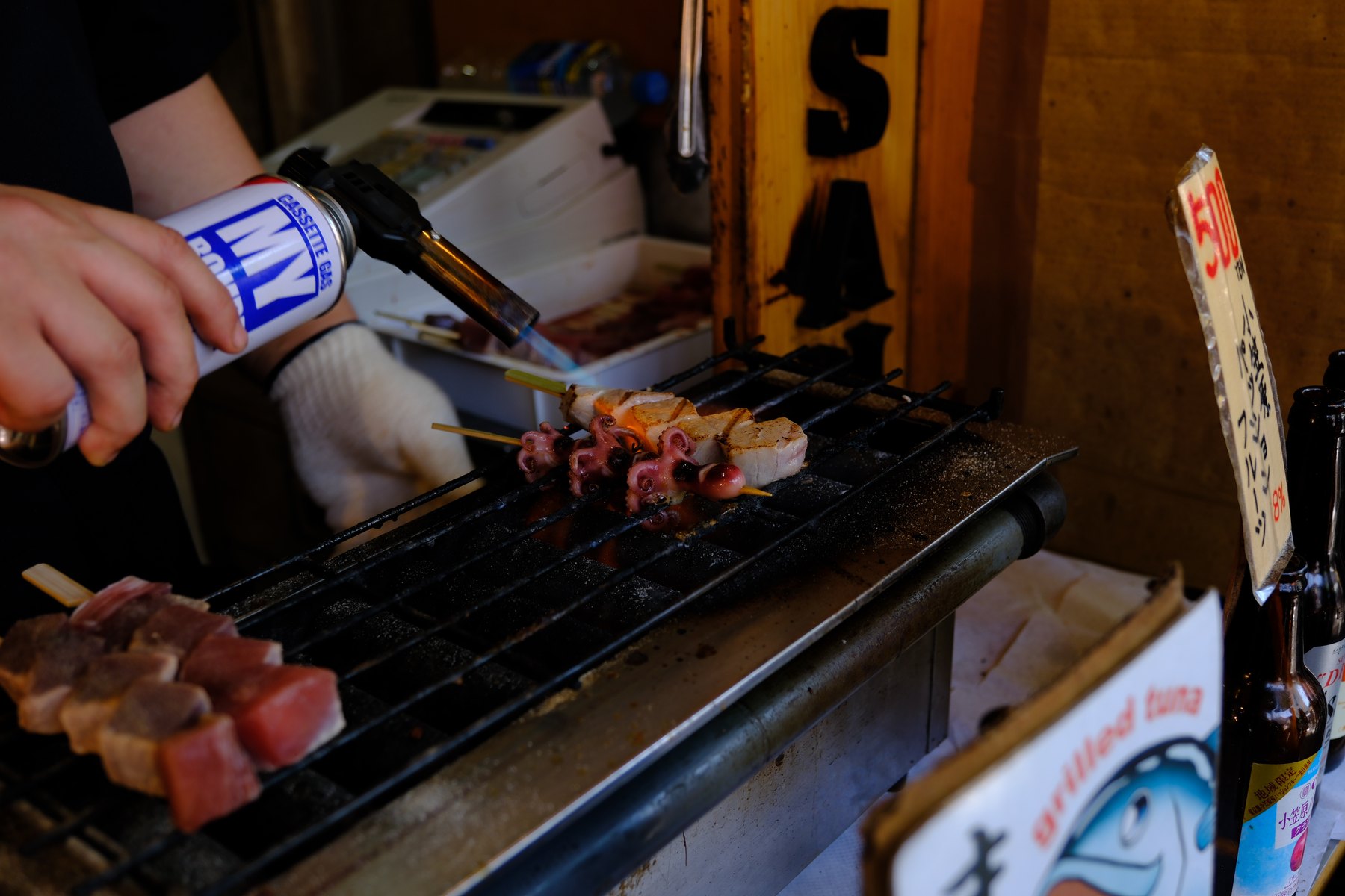A street-food vendor using a torch over skewers at a Tokyo stall, with no face visible